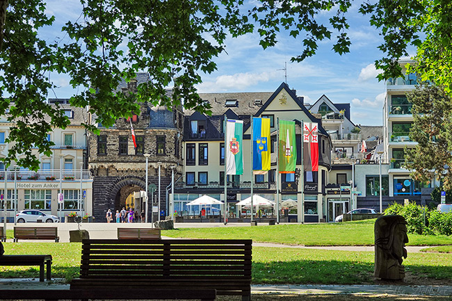 Blick durch grüne Blätter auf das historische Rheintor in Andernach. Davor eine Wiese und die Rheinpromenade mit dem neu gestalteten Außenbereich eines Hotels mit weißen Sonnenschirmen.