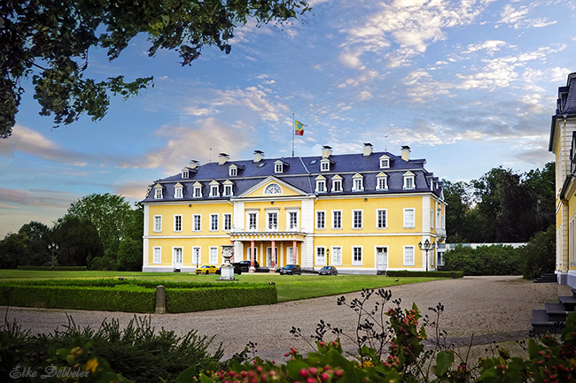 Frontansicht des gelben, barocken Schloss Neuwied an einem sonnigen Tag, umgeben von einem gepflegten Park mit grünen Rasenflächen und Bäumen unter einem leicht bewölkten Himmel.