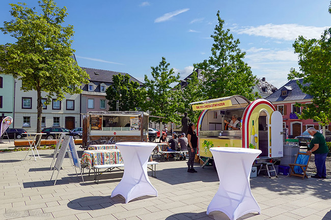 Zwei weiße Stehtische im Vordergrund auf dem Feierabendmarkt, im Hintergrund sind Foodtrucks und Sitzgelegenheiten zu sehen.