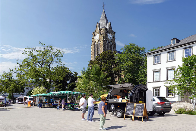 Gesamtansicht des Feierabendmarktes in Neuwied mit Marktständen, einigen Besuchern und der Marktkirche im Hintergrund unter blauem Himmel.