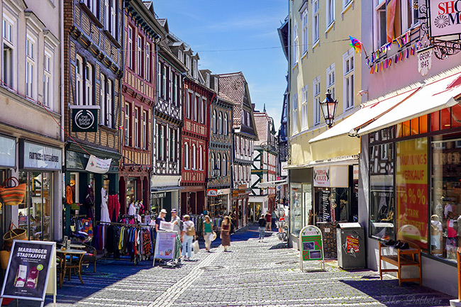  Eine belebte Kopfsteinpflasterstraße in Marburg, gesäumt von mehrstöckigen, bunten Fachwerkhäusern mit Geschäften im Erdgeschoss. Menschen spazieren auf dem Bürgersteig, Sonnenschein erhellt die Szene.  Weitere Fotos für diesen Artikel: