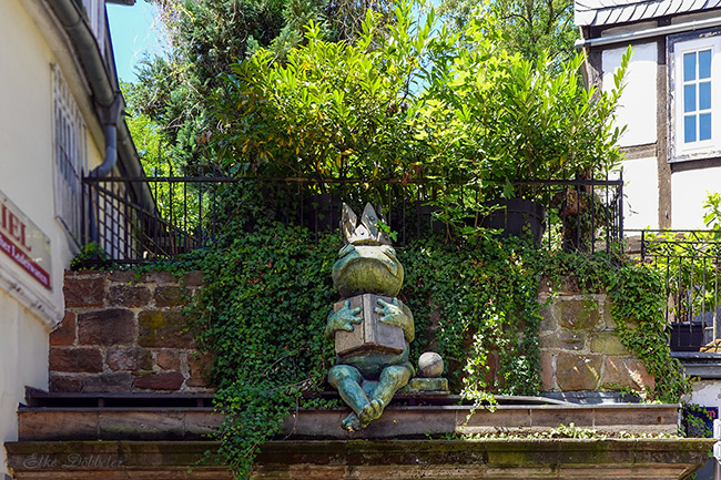 Eine kleine Bronze-Skulptur eines Frosches mit Krone, der auf einem SIms sitzt.