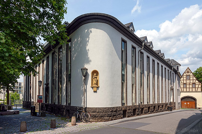Helles, langgestrecktes Gebäude in Andernach mit abgerundeter Ecke und schmalen, hohen Fenstern. An der Fassade befindet sich eine kleine Nischenfigur. Das Haus steht an einer ruhigen Straße mit Kopfsteinpflaster, Bäumen und blauem Himmel mit weißen Wolken im Sommer.