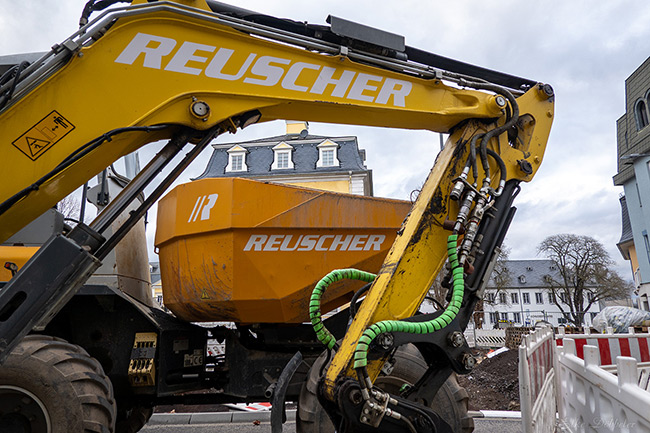 Ein großer gelber Hydraulikbagger der Firma Reuscher steht still auf der Baustelle in der Schlossstraße vor den Fassaden historischer Gebäude.