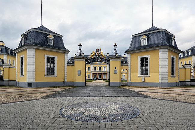 Blick auf das gelbe Schloss Neuwied durch das schmiedeeiserne Haupttor mit goldenem Wappen. Im Vordergrund der neu gepflasterte Schlossplatz mit einem kreisrunden Mosaik, das einen hellen Pfau auf dunklerem Grund darstellt. Der Himmel ist bewölkt.
