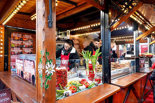 Ein rustikaler Holzstand auf dem Currywurstfestival. Im Vordergrund stehen Gläser mit roten Chilis und Frühlingszwiebeln, dahinter bereitet das Personal frische Würste auf dem Grill zu. Foto: Elke Döbbeler