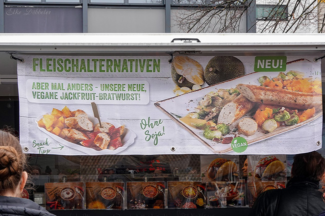 Eine Werbebanner an einem Imbisswagen für Fleischalternativen. Zu sehen ist eine neue, vegane Jackfruit-Bratwurst ohne Soja, serviert mit Beilagen. Foto: Elke Döbbeler