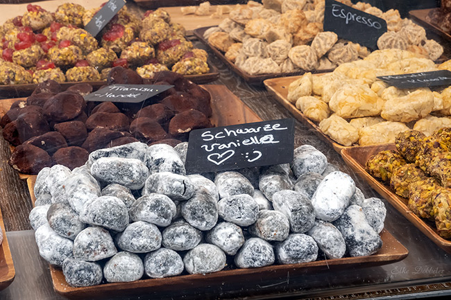 Eine Auslage mit verschiedenen italienischen Gebäckstücken. Im Fokus stehen dunkle Kugeln mit Puderzucker, die als "Schwarze Vanille" ausgeschildert sind, daneben Sorten wie Tiramisu-Mandel und Espresso. Foto: Elke Döbbeler