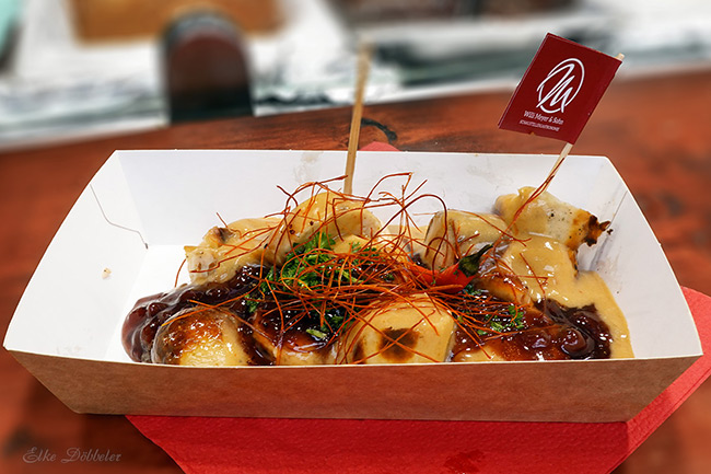 Eine Nahaufnahme einer Currywurst in einer Pappschale, garniert mit feinen roten Chilifäden, Kräutern und zwei verschiedenen Saucen. Ein kleiner Deko-Stecker steckt im Gericht. Foto: Elke Döbbeler