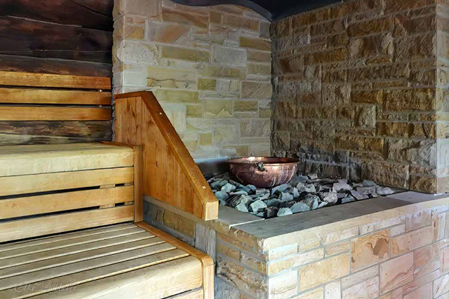 Nahaufnahme eines Saunaofens mit Natursteinwand, Saunasteinen und einer Kupferschale für Aufgüsse neben hölzernen Sitzbänken.