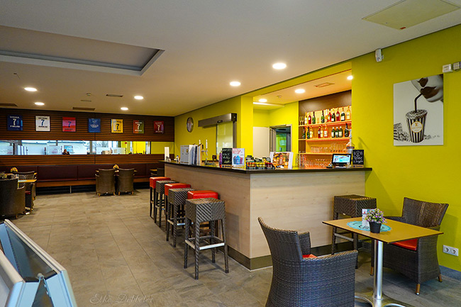 Moderner Bistrobereich der Saunalandschaft mit einer hellen Theke, Barhockern und kleinen Tischen vor einer leuchtend gelben Wand.