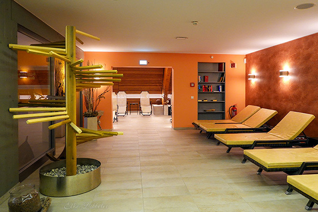 Warmer Ruheraum mit gelben Entspannungsliegen, rötlich gestalteten Wänden und einer dekorativen Holzskulptur in Baumform.