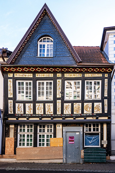 Nahaufnahme eines historischen Fachwerkhauses in Ahrweiler mit dunkelblauem Schieferdach und kunstvollen Schnitzereien, die Weintrauben und Reben zeigen. Im unteren Bereich sind Infotafeln zur Stadtgeschichte angebracht.