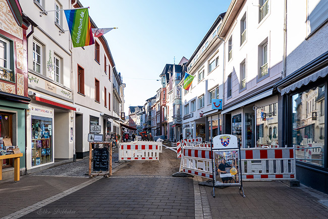 Eine Straßenszene in der Fußgängerzone von Ahrweiler. Man sieht helle Häuserfassaden mit bunten „wiederbunt.de“-Flaggen und rot-weiße Baustellenabsperrungen, die auf die laufenden Pflasterarbeiten hinweisen.