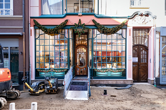 Frontalansicht einer aufwendig restaurierten Jugendstil-Fassade mit grünen Fensterrahmen und festlicher Beleuchtung. Unmittelbar davor liegt eine unbefestigte Erdbaustelle mit zwei Rüttelplatten und einem Bagger-Kettenlaufwerk im Vordergrund.