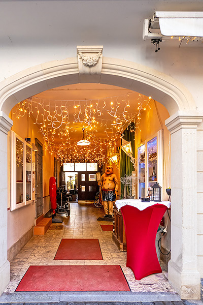 Ein heller Torbogen, der in einen mit warmen Lichterketten geschmückten Gang führt. Im Hintergrund sieht man eine Koch-Figur und einladende Dekoration.