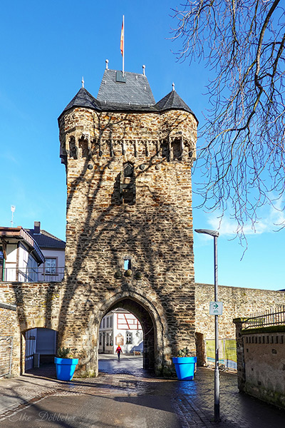 Ein massives, mittelalterliches Stadttor aus Bruchstein mit zwei blauen Blumenkübeln davor. Durch den Torbogen blickt man in eine helle Gasse mit Fachwerkhäusern unter strahlend blauem Himmel.