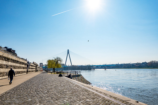 Weitwinkelaufnahme des Rheinufers in Neuwied an einem sonnigen Tag. Im Vordergrund ein gepflasterter Uferweg mit einer hohen Steinmauer links und einem Spaziergänger. Im Hintergrund die markante Raiffeisenbrücke unter einem strahlend blauen Himmel mit einer hellen Sonne oben rechts.