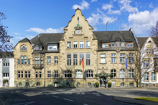 Historische Fassade des Kreishauses Neuwied am Moltkeplatz mit Sandsteingliederung, geschwungenem Giebel, Uhrturm und zwei wehenden Flaggen (Deutschland und Europa) bei blauem Himmel. Vor dem Gebäude ist der Eingangsbereich mit einem modernen Glasvordach zu sehen.