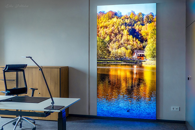 Ein helles Büro in der Sparkasse Neuwied. Im Fokus steht ein raumhohes, von innen beleuchtetes Wandbild. Es zeigt den Schwanenteich in Oberbieber mit leuchtend gelben und orangefarbenen Herbstbäumen, die sich im blauen Wasser spiegeln. Links im Vordergrund sieht man einen modernen Schreibtisch und einen Bürostuhl.