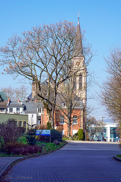 Blick von der gepflasterten Auffahrt des Marienhaus Klinikums St. Elisabeth in Neuwied auf den hohen, markanten Kirchturm der Sankt-Matthias-Kirche unter strahlend blauem Himmel. Im Vordergrund sind Bäume mit ersten Frühlingsknospen und die moderne Architektur des Klinikgeländes zu sehen.
