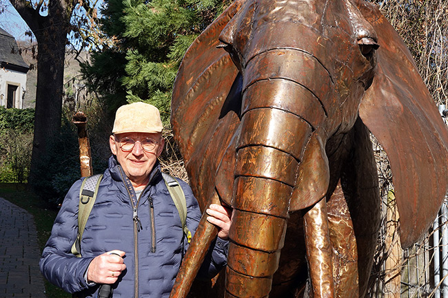 Willi steht lächelnd neben einer großen, rostfarbenen Elefantenskulptur im Skulpturenpark. Er trägt eine beige Kappe und einen Rucksack.