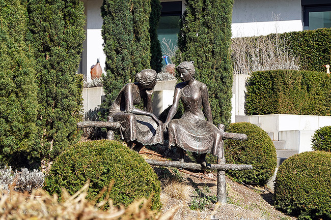 Bronzeskulptur „Zwiegespräch“ von Jürgen Ebert: Zwei lebensgroße Frauenfiguren sitzen entspannt auf einem Holzzaun und unterhalten sich im Skulpturenpark Andernach.